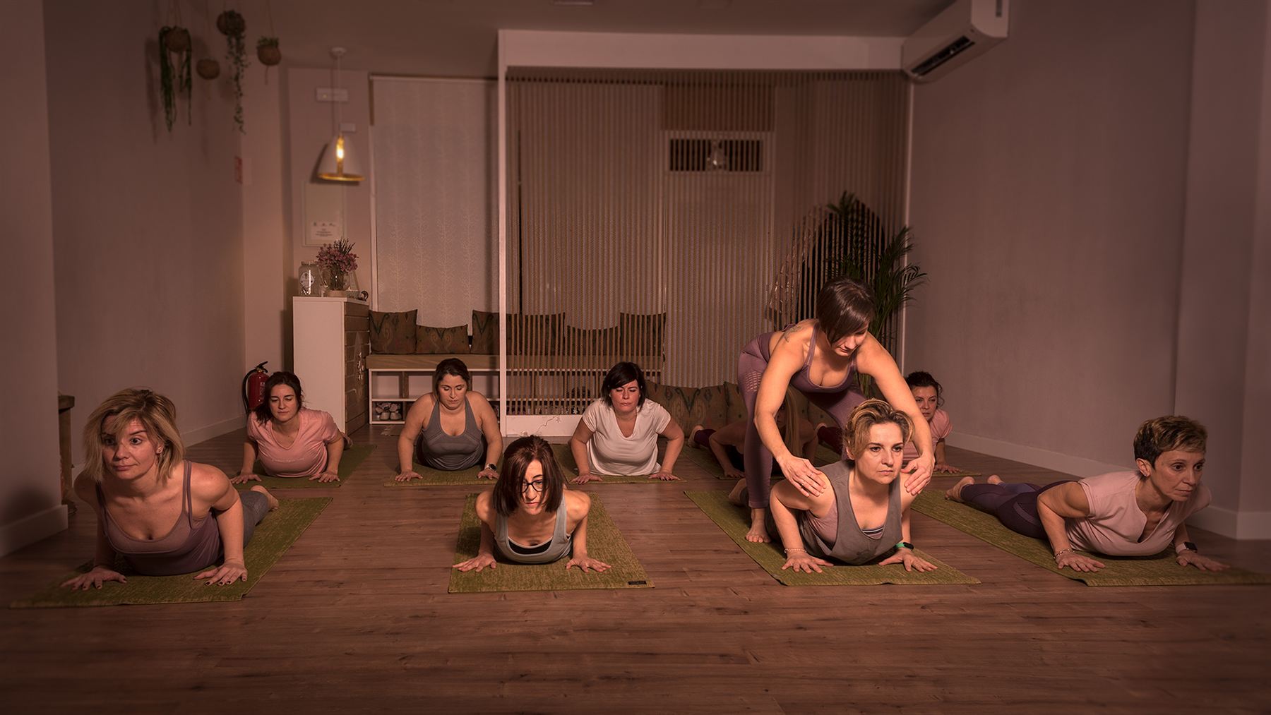 Grupo de alumnas en la postura de la cobra mientras Gema ajusta con las manos los hombros de una de ellas, en la sala de práctica de luz cálida.